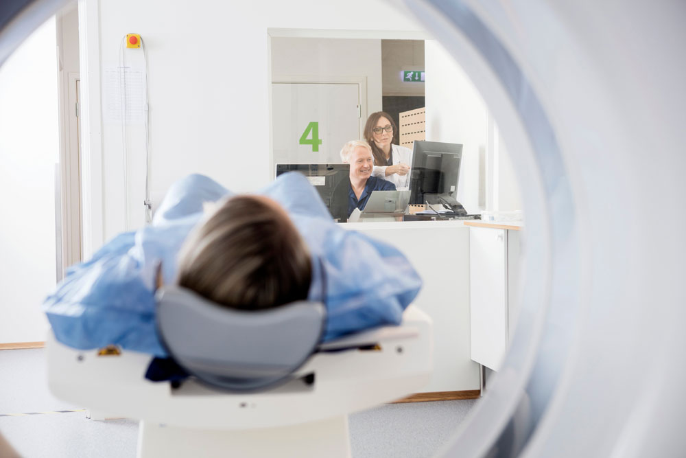 in the foreground, a patient undergoes a CT scan, while in the background, two doctors discuss the image on a computer screen. The view is from behind the patient's head, looking out of the CT scanner toward the tech booth where the doctors sit.